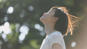 木漏れ日の差す屋外で目を閉じる女性。花粉症やアレルギー性結膜炎による目のかゆみ対策を紹介する記事のイメージ写真。